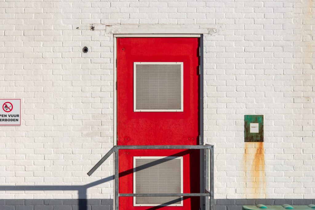 Bright red industrial door with warning sign on white brick wall, sunlight casting shadows.